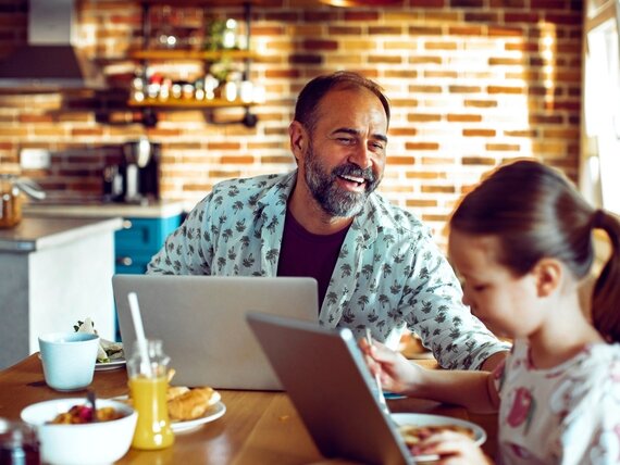 Ein Vater sitzt mit seiner Tochter in der Küche beim Frühstück, während am Laptop arbeitet.
