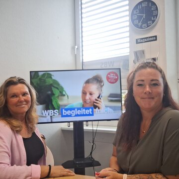 Beratungsteam mit viel Herz. 2 Frauen sitzen in einem Büro vor einem Bildschirm und blicken freundlich in die Kamera.