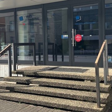 Vierstufige Treppe zum Haupteingang. Glastüre mit WBS TRAINING Logo und blauer Aufschrift „Herzlich Willkommen“.