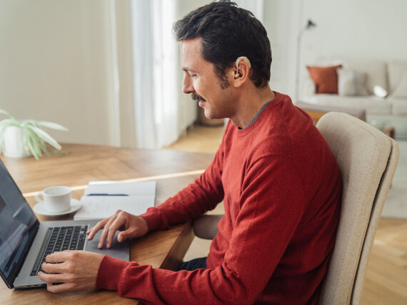 Ein Mann mit Hörgerät sitzt fröhlich vor seinem Laptop.