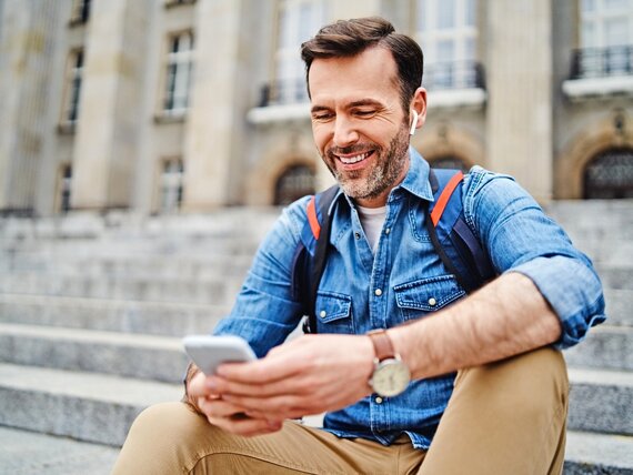 Ein Mann mit Kopfhörern in den Ohren sitzt draußen auf einer Treppe und schaut auf sein Smartphone.