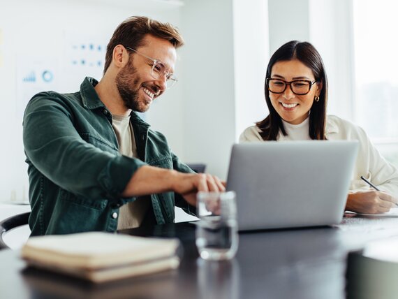 Ein gut gelaunter Mann zeigt seiner Kollegin etwas an seinem Laptop.