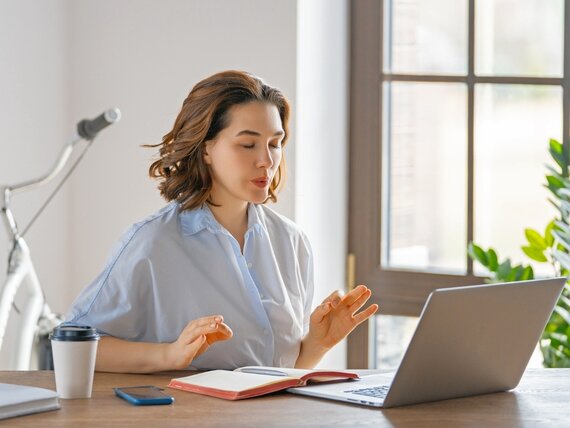 Eine junge Frau sitzt vor ihrem Laptop und macht Atemübungen mit geschlossenen Augen.