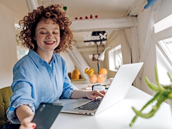 Eine Frau im Homeoffice arbeitet am Laptop und schaut auf ihr Smartphone.