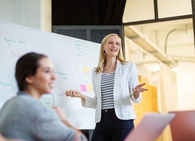 Eine junge, selbstbewusste Frau steht an einem Whiteboard und erklärt ihrem Team ihre Ideen