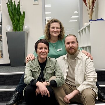 Nahaufnahme von 3 Personen, die auf einer Treppe sitzen und freundlich in die Kamera lächeln.