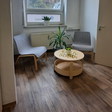 Kleiner Pausenraum mit 2 Sesseln und einem Beistelltisch mit Deko. Im Hintergrund ein großes Fenster und eine Zimmerpflanze.