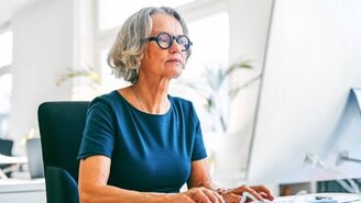 Eine Frau sitzt am Schreibtisch und schaut auf einen Bildschirm. Hände auf der Tastatur.