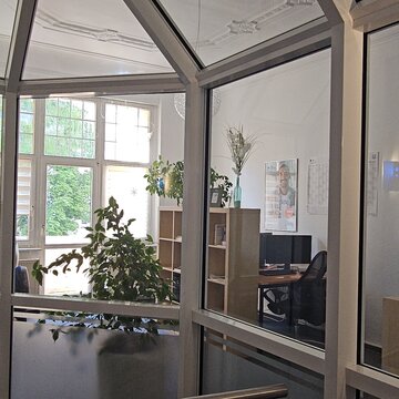 Blick durch Glaswand auf einen Schulungsraum mit mehreren Arbeitsplätzen. Mittig ein großes Regal und verteilt im Raum mehrere Pflanzen.