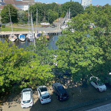 Sicht aus dem Fenster am Standort. Ein Gewässer mit Segelbooten und Bäume sowie einige geparkte Autos.