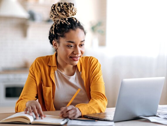 Eine Frau sitzt in der Küche vor ihrem Laptop und macht sich Notizen.