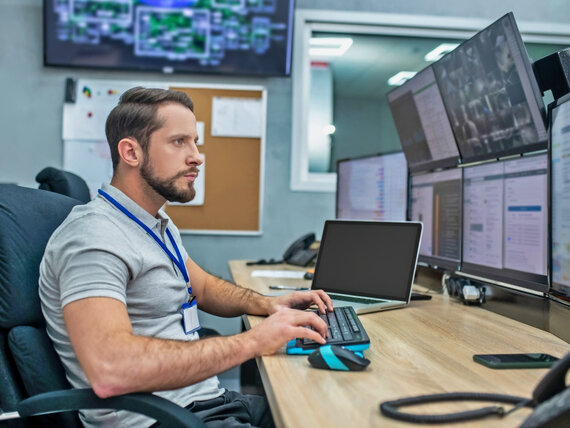 Eine männliche Wach- und Sicherheitsfachkraft sitzt vor mehreren Monitoren und überwacht eine Gebäudeanlage