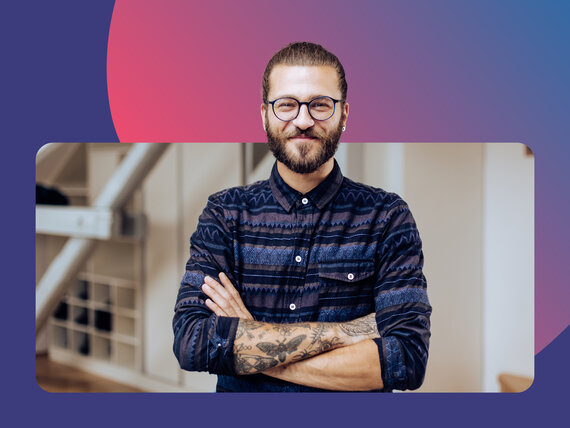 Mann mit Brille, Bart und Tattoo steht mit verschränkten Armen in einem Raum und lächelt