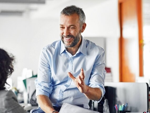 Ein Mann sitzt locker auf einem Schreibtisch im Büro und unterhält sich mit seiner Kollegin.