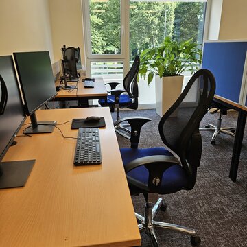 Teilnehmerraum mit mehreren Arbeitsplätzen, jeweils mit zwei Bildschirmen, Bürostuhl, Tastatur, Maus