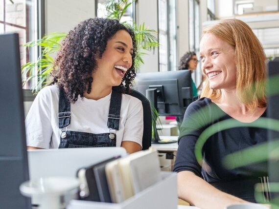 Zwei Frauen sitzen im Büro an ihren Computern und lachen fröhlich.
