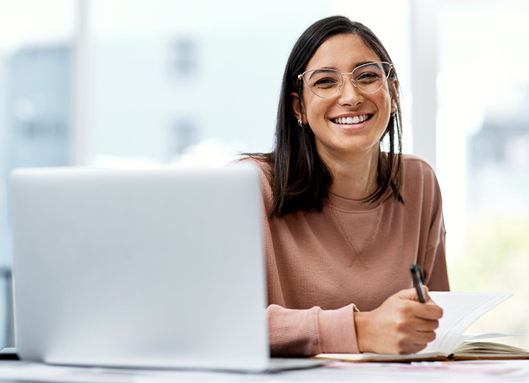 Junge Frau mit Brille sitzt in einem hellen Buero am Laptop und lacht froehlich in die Kamera.