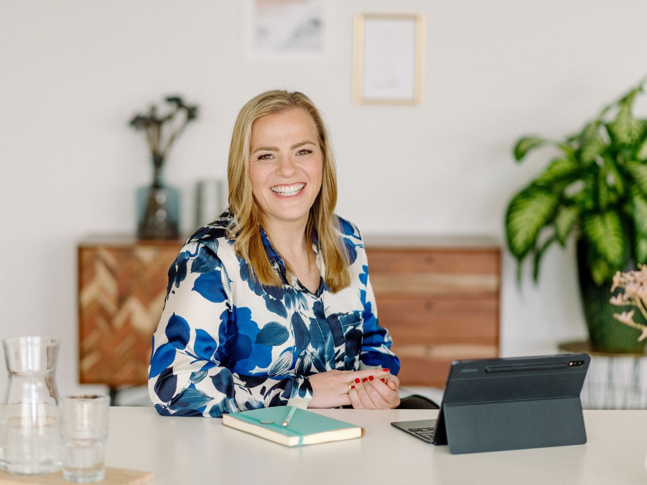 Frau mit langen Haaren und blau‑geblümtem Kleid sitzt lächelnd an einem Tisch mit Tablet und Notizbuch.