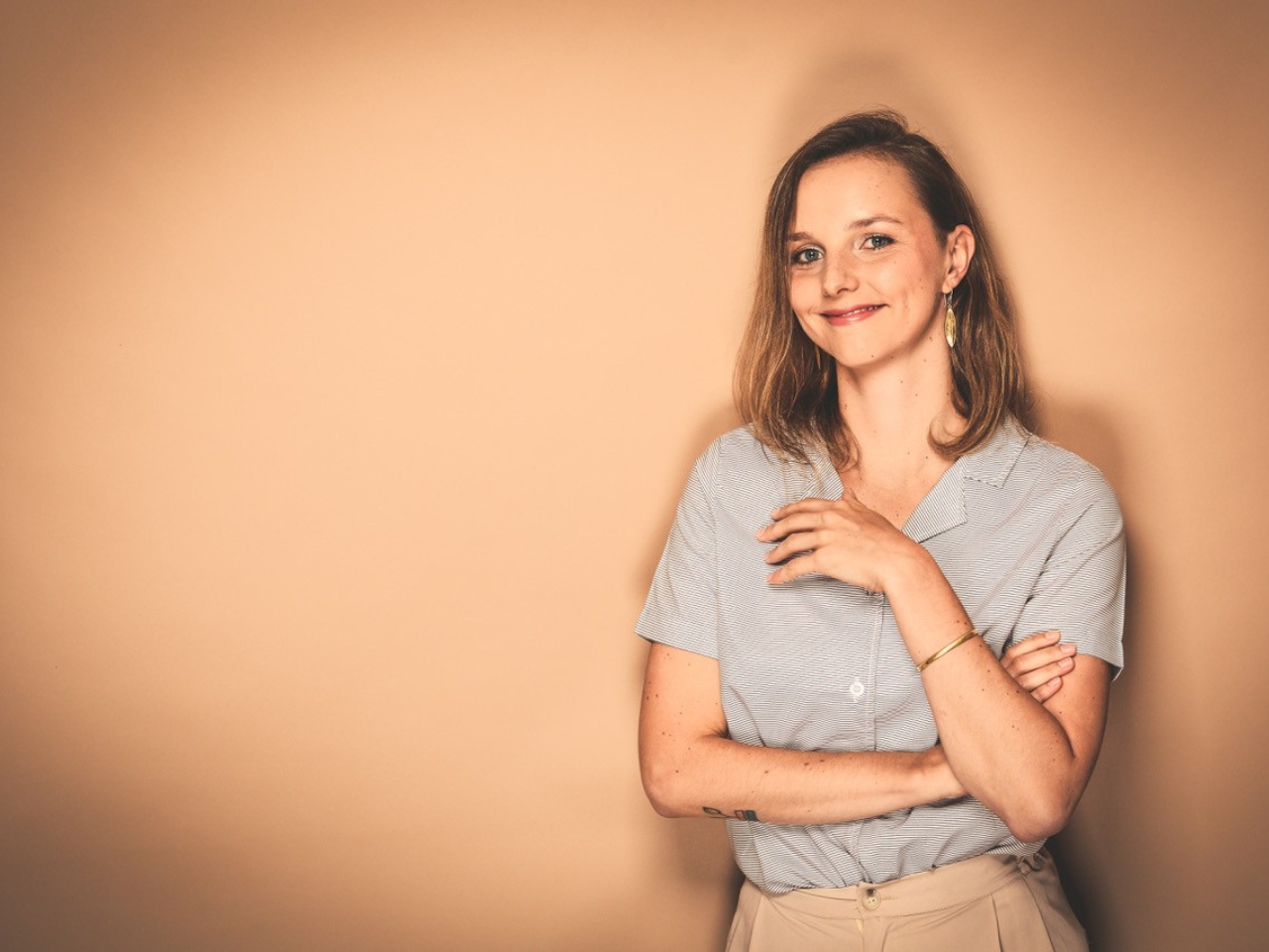 Eine Frau mit grauer Bluse steht vor einer orangefarbenen Wand und lächelt in die Kamera.